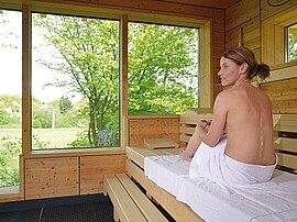Frau sitzt in der Sauna und schaut durch ein Fenster in die grüne Landschaft während ihres Wellnessurlaubs