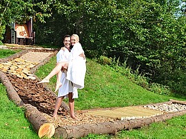 Mann im Bademantel hebt Frau im Bademantel während des Saarland Urlaubs auf dem Barfußpfad hoch. 